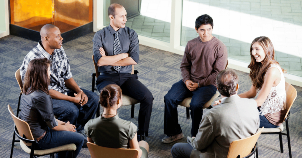 Team of seven people sitting in a circle on chairs having a meeting.