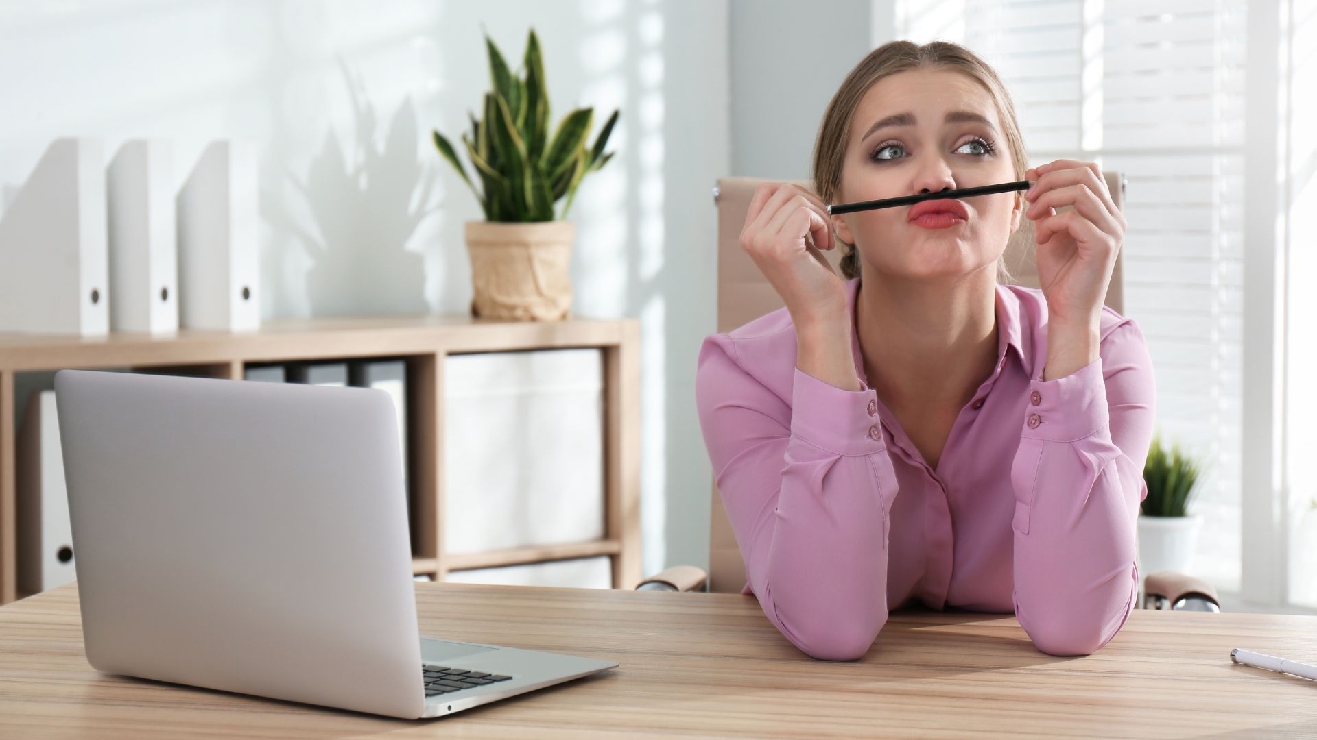 Women sitting at desk with laptop, has a pen between her mouth and nose